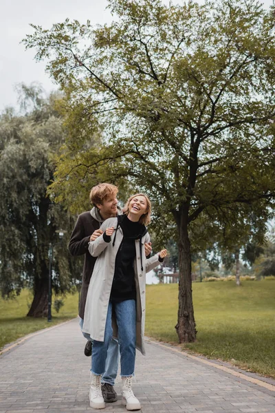 Homme heureux en manteau étreignant jeune femme joyeuse avec les yeux fermés riant dans le parc — Photo de stock