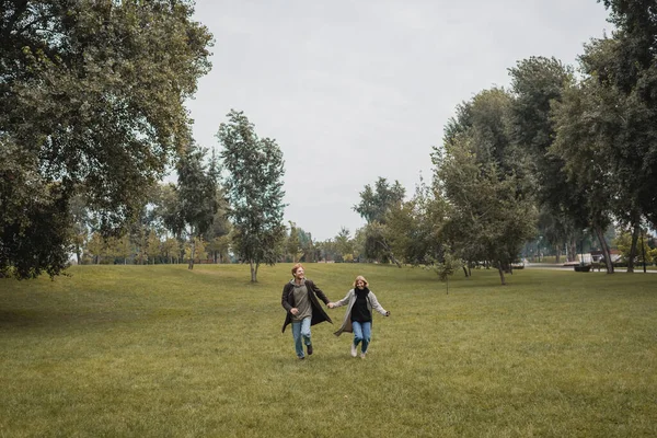 Full length of happy man and pleased woman in coats holding hands while running on grass — Stock Photo