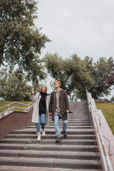 Cheerful blonde woman and redhead man with umbrella holding hands while descending stairs in park — Stock Photo