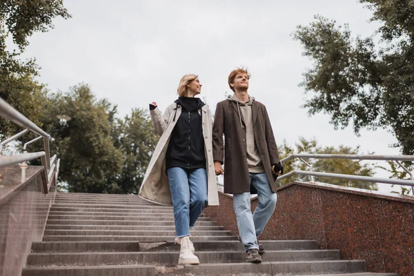 Low angle view of cheerful woman and redhead man with umbrella holding hands while descending stairs near park — Stock Photo