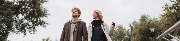 Low angle view of cheerful woman and redhead man against cloudy sky in park, banner — Stock Photo