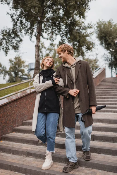Femme heureuse et rousse homme avec parapluie descendant escaliers dans le parc — Photo de stock