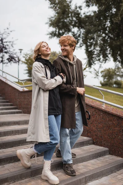 Joyful woman and happy redhead man with umbrella descending stairs in park — Stock Photo