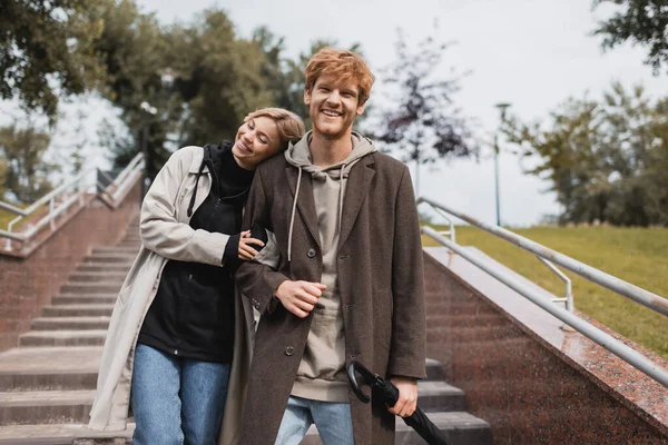 Pleased woman leaning on shoulder of redhead man with umbrella while descending stairs in park — Stock Photo