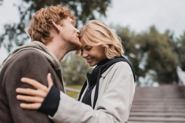 Ruiva homem beijar testa de feliz namorada no parque — Fotografia de Stock