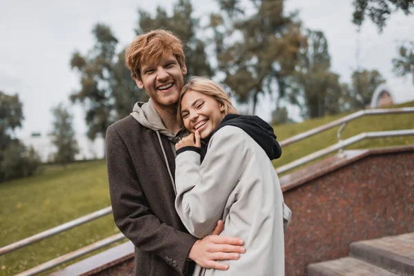 Happy young woman hugging with joyful redhead boyfriend in autumnal coat in park — Stock Photo