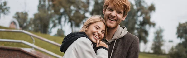 Happy young woman hugging redhead boyfriend in autumnal coat in park, banner — Stock Photo