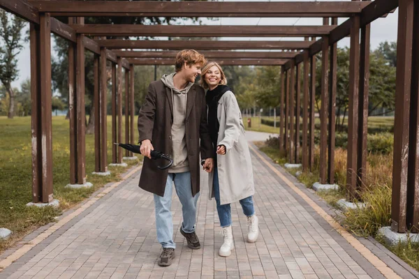 Pleine longueur de heureux jeune couple en manteaux automnaux tenant la main tout en marchant sous l'arc multiple dans le parc — Photo de stock