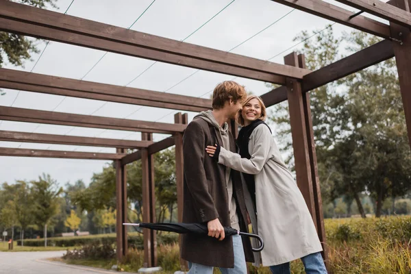 Усміхнена молода пара в акумуляторних пальто обіймається під декількома арками в парку — стокове фото