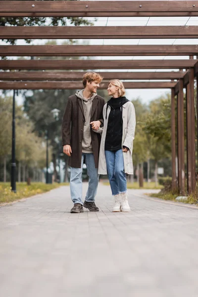 Повна довжина щасливої пари в автентичних пальто тримає руки під час прогулянки під декількома арками в парку — стокове фото
