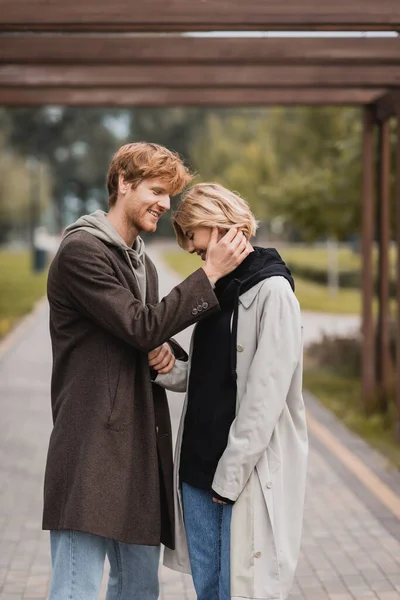 Heureux jeune homme en manteau automnal étreignant femme sous arche dans le parc — Photo de stock