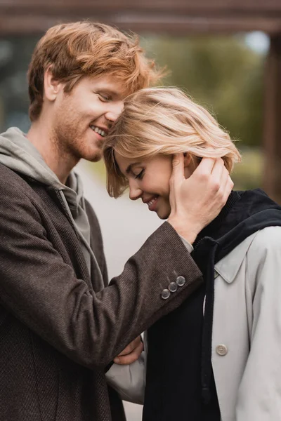 Feliz joven en abrigo otoñal abrazando novia rubia con los ojos cerrados - foto de stock