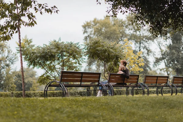 Alegre joven hombre y mujer abrazando y sentado en banco de madera en el parque verde - foto de stock