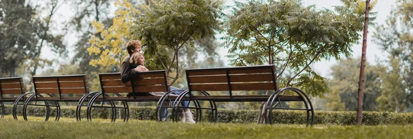 Alegre joven hombre y mujer abrazando y sentado en el banco de madera en el parque verde, pancarta - foto de stock