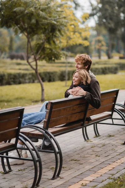 Веселый молодой человек обнимает женщину и сидит на деревянной скамейке в парке — стоковое фото