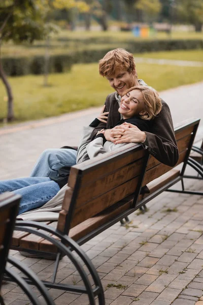 Cheerful young man hugging positive woman and sitting on wooden bench in park — Stock Photo