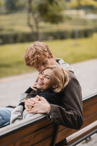 Pelirroja hombre abrazando y besando a mujer positiva mientras está sentado en banco de madera en el parque - foto de stock