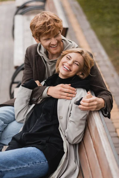 Vista ad alto angolo di allegro giovane uomo che abbraccia donna sorridente e seduta su una panchina di legno nel parco — Foto stock