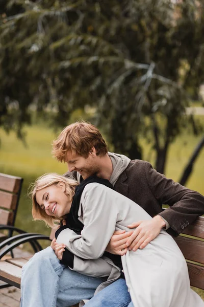 Рыжий мужчина смеется, обнимая счастливую девушку, сидя на скамейке — стоковое фото