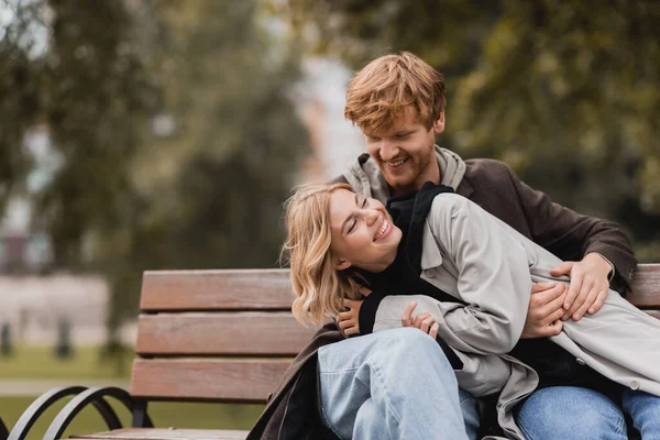 Rossa uomo sorridente mentre abbraccia allegra fidanzata mentre seduto su panchina — Foto stock