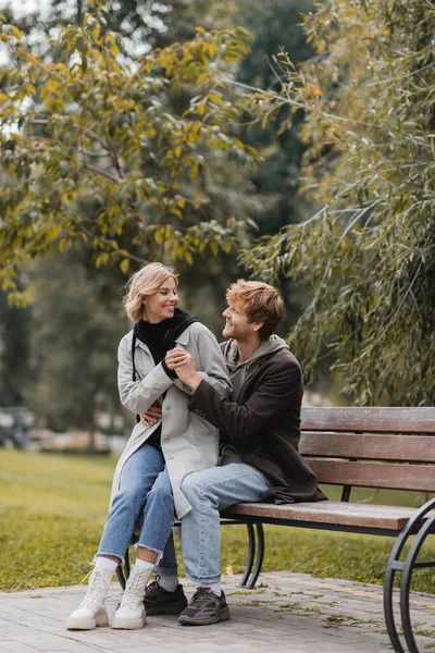 Pelirroja hombre sonriendo mientras abraza a novia positiva mientras está sentado en el banco - foto de stock