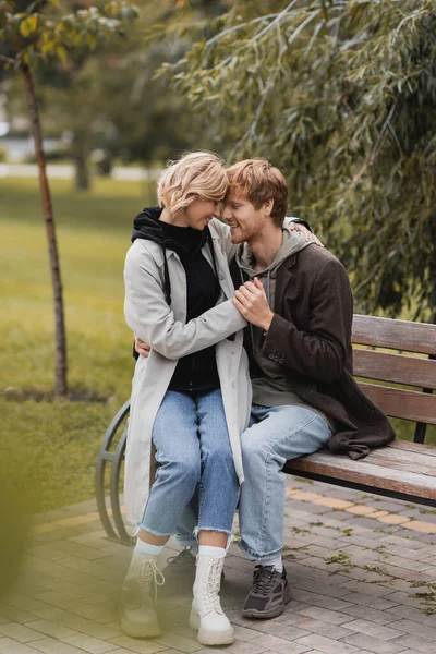 Pelirroja hombre sonriendo mientras toma de la mano con su novia positiva mientras está sentado en el banco - foto de stock