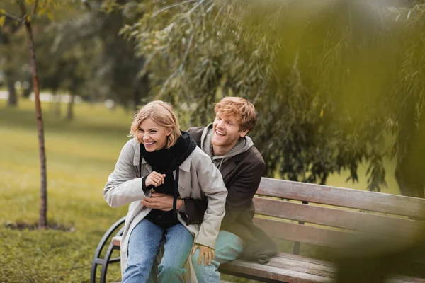 Redhead man smiling while tickling positive girlfriend while sitting on bench — Stock Photo