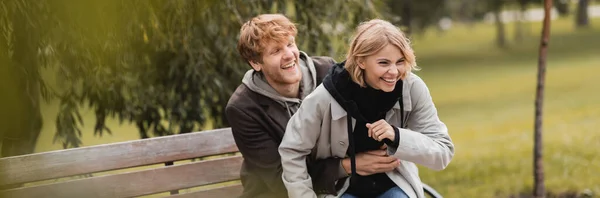 Rothaarige Mann lächelt, während kitzelt positive Freundin in der Nähe Bank im Park, Banner — Stockfoto