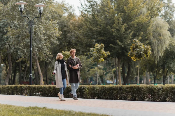 Full length of redhead man and blonde woman in coat smiling while walking in park — Stock Photo
