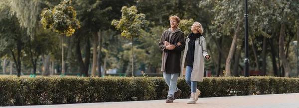 Volle Länge des rothaarigen Mannes und der blonden Frau im Mantel, die beim Spazierengehen im Park lächelt, Banner — Stockfoto