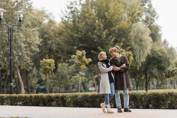 Longitud completa de hombre pelirrojo y mujer rubia en abrigo sonriendo mientras se miran en el parque - foto de stock