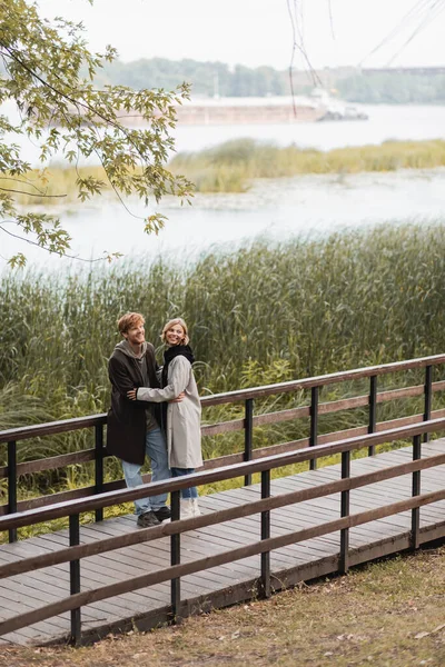 Comprimento total do homem ruivo e mulher feliz no casaco sorrindo ao abraçar na ponte perto do lago no parque — Fotografia de Stock