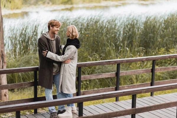 Pleine longueur de rousse homme et femme blonde en manteau souriant tout en étreignant sur le pont près du lac dans le parc — Photo de stock