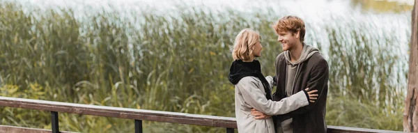 Rothaariger Mann und blonde Frau im Mantel lächeln, während sie sich auf Brücke am See im Park umarmen, Banner — Stockfoto