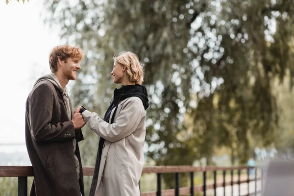 Ruiva homem e mulher loira no casaco sorrindo enquanto segurando as mãos perto da ponte no parque — Fotografia de Stock