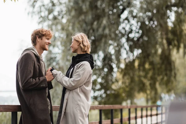 Rossa uomo e bionda donna in cappotto sorridente mentre si tiene per mano vicino al ponte nel parco — Foto stock