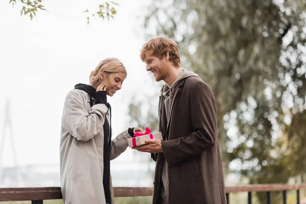 Happy redhead man holding wrapped present near cheerful girlfriend in park — Stock Photo
