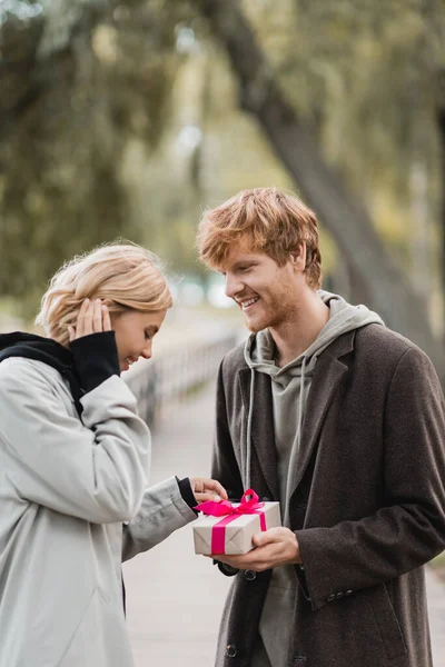 Feliz pelirroja hombre sosteniendo envuelto presente cerca complacido novia en parque - foto de stock