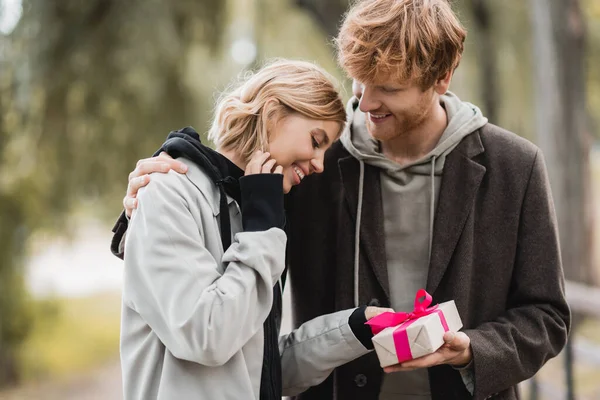 Feliz pelirroja hombre celebración envuelto caja de regalo cerca complacido novia en parque - foto de stock