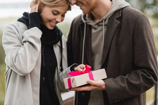 Happy man holding wrapped gift box with pink ribbon near pleased girlfriend in park — Stock Photo