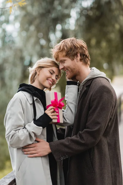 Happy woman holding wrapped gift box with pink ribbon near pleased boyfriend in park — Stock Photo