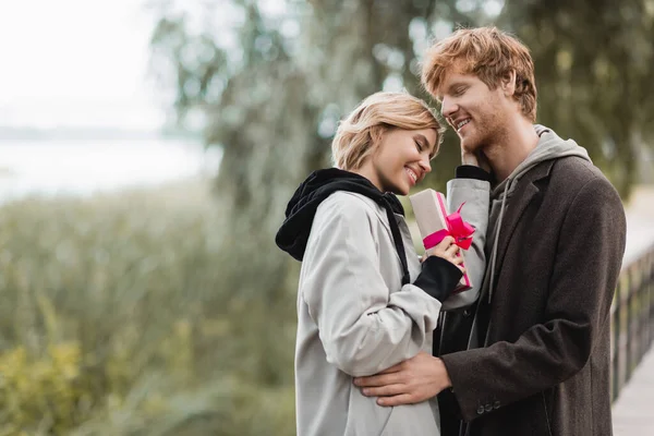 Glückliche Frau hält verpackte Geschenkschachtel mit rosa Schleife neben fröhlichem Freund im Park — Stockfoto