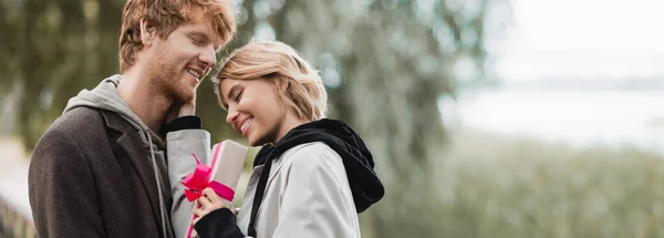 Happy woman holding wrapped gift box with pink ribbon near smiling boyfriend in park, banner — Stock Photo