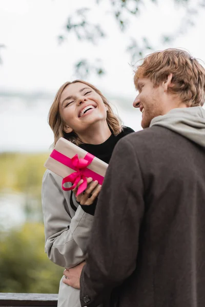 Happy woman laughing while holding wrapped gift box near cheerful boyfriend in park — Stock Photo