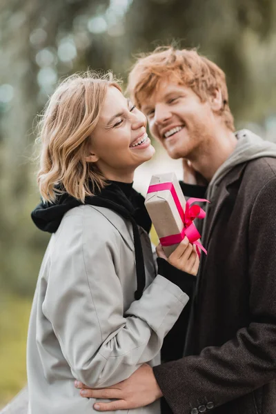 Happy woman laughing while holding wrapped gift box near cheerful boyfriend on blurred background — Stock Photo
