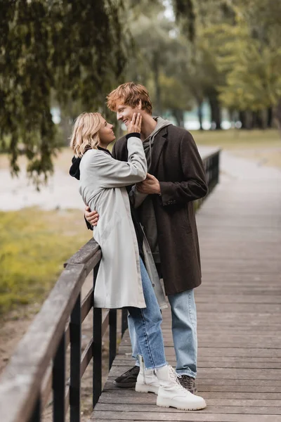 Рыжеволосый мужчина и блондинка в пальто обнимаются во время свидания в парке — стоковое фото
