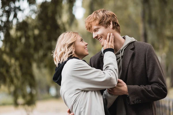 Redhead man and blonde woman in coat smiling while having date in park — Stock Photo