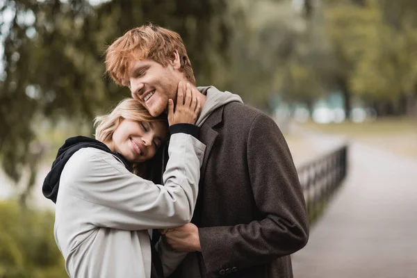 Довольный рыжий мужчина и блондинка в пальто улыбается во время свидания в парке — стоковое фото