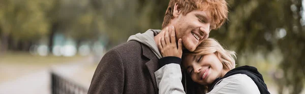 Hombre pelirrojo complacido y mujer rubia en abrigo sonriendo mientras tiene fecha en el parque, pancarta - foto de stock