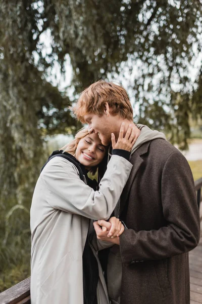 Alegre pelirroja hombre y complacido mujer en abrigo sonriendo mientras tener fecha en parque - foto de stock